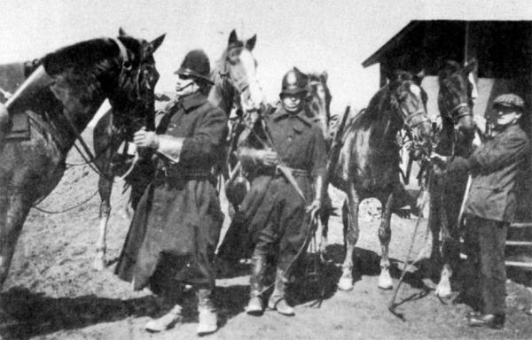 Two policemen stand next to their horses, while a stable person holds two horses by the reins.
