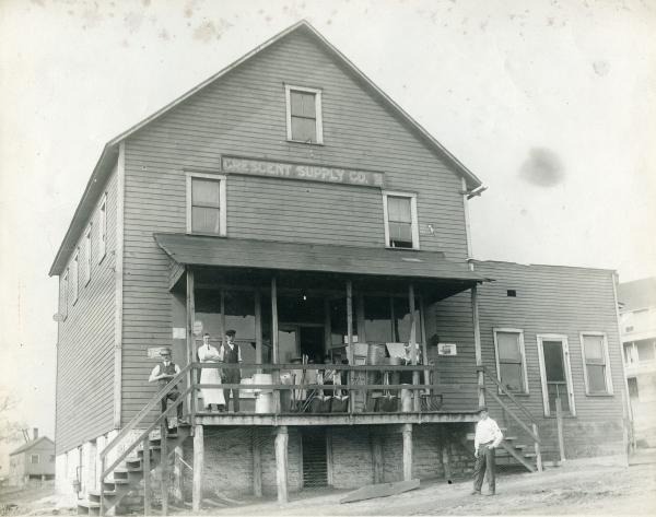 A black and white photograph of a wooden company store with a large porch. Two employees and several small children stand on the porch. One employee stands on the steps extending from the left of the porch. One man stands in front of the store, to the right of the photograph. There is a sign above two windows that reads: Crescent Supply Co.