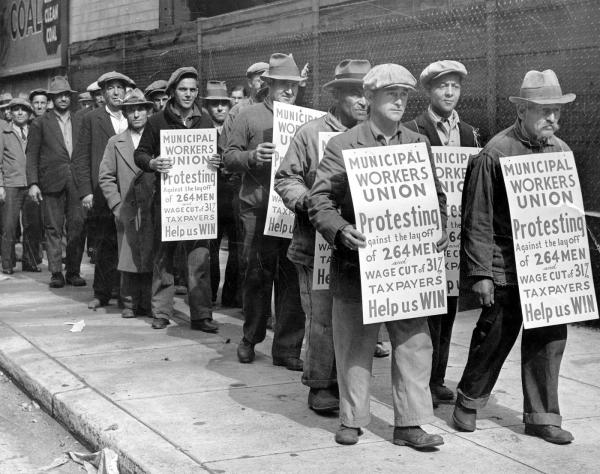 Marching strikers wear placards that read Municipal Workers Union protesting against the Layoff of 264 Men and wage cut of 31% Taxpayers Help Us Win.