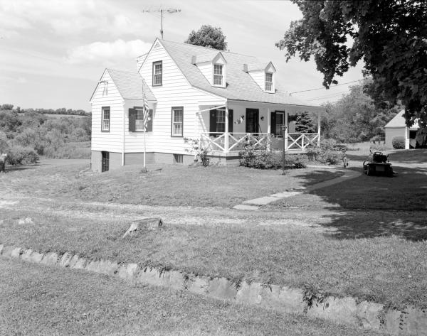Black and white photograph of the exterior of a Norvelt House. A curved foot path leads to a white clapboard two-story house with a porch. A flag pole and a riding lawn mower rest outside. 