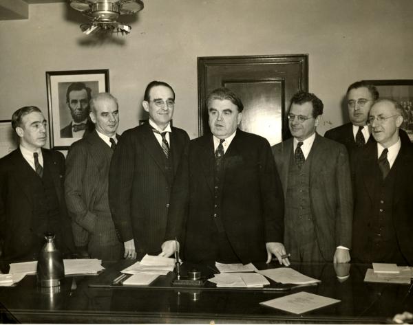 Black and white press photograph of seven men dressed in suits, standing behind a desk. On the wall, in the upper left hand corner of the photograph is a picture of Abraham Lincoln.