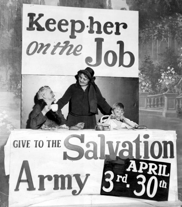 An elderly lady sits holding a cup, a seated, young boy, takes a bite of food. A salvation army worker stands between them with outstretched arms, her hands touching a shoulder of each. All are inside of a booth that has two signs. One sign reads: Keep her on the Job and the other reads: Give to the Salvation Army, April 3rd 30th .