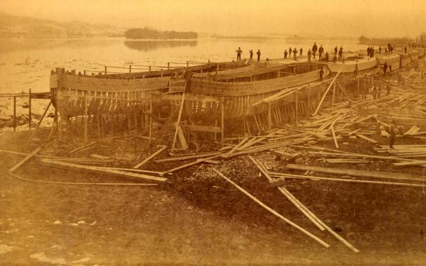 Workers stand along the tops of two long rows of canal boats and along the side of wooden scaffolding that holds the boats that are in process of being constructed.  