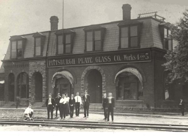 Black and white image of the exterior of a building with arched windows and doors. A group of men and women pose for this photo, in front of the shop and behind the railroad tracks.