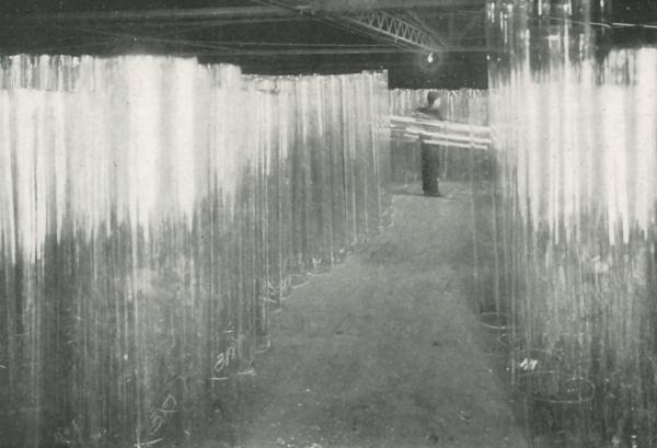An interior view of a glass factory full of storied window glass and in the background a worker can be seen standing holding a cylinder.