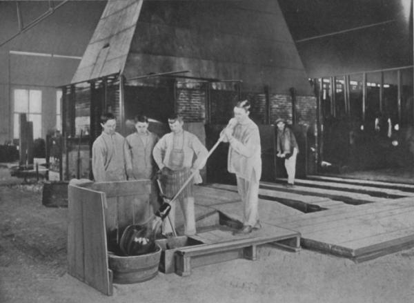 A glass blower works with a blowing iron and molten glass to blow a window-glass cylinder. Other workers are observing the task.