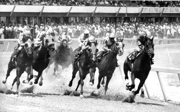 Jockeys on their horses round a corner of a race track, dirt flies in the air, and a crowd of spectators watch.