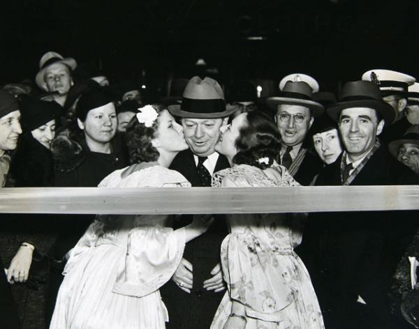 Philadelphia Mayor S. Davis Wilson being greeted at the opening of a new Florsheim Show Store. At the entrance of a ribbon cutting, two women kiss the face cheeks of a man in a suit. 