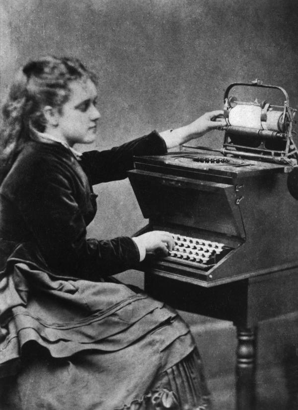 A young lady, wearing a long skirt and a long sleeve jacket, sits at a typewriter.
