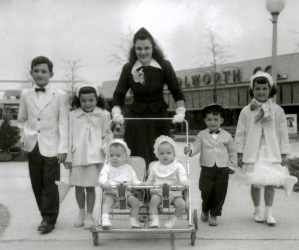 Image of young mother pushing a stroller carrying twins, while two young boys and two young girls dressed in their Easter clothes walk beside her. Woolworth's can be seen in the background.