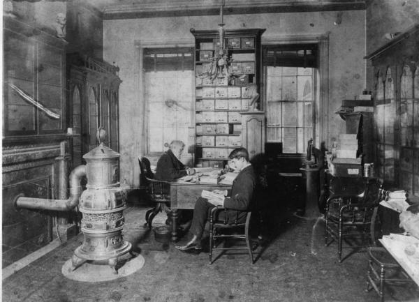 Two men working in an office, one seated at a desk and one seated in a chair next to the desk. A huge pot belly stove is in the room.
