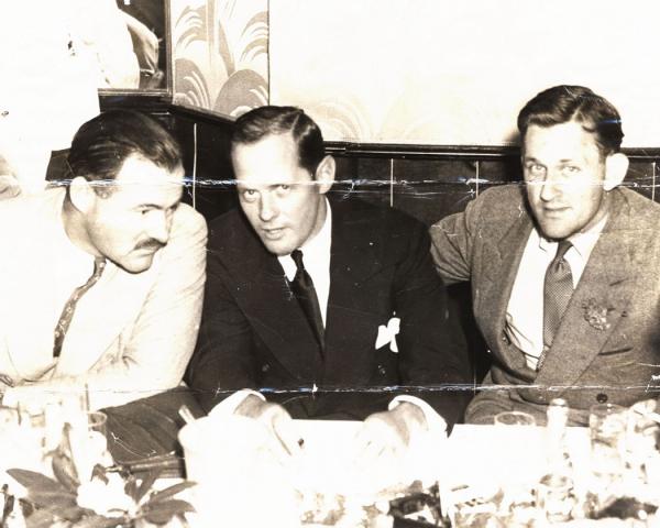 Three men sit at a table. Let to right, Hemingway, Billingsly, and O'Hara