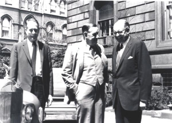 Black and white photograph of three men, standing on a city sidewalk, conversing.