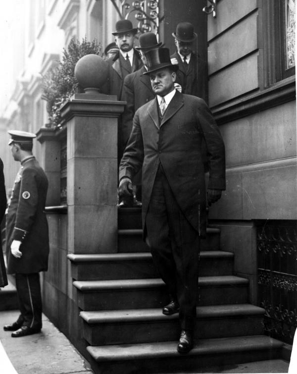 Tener exiting a building, walking down a fight of stairs, dressed in a tuxedo and top hat. Several men follow behind him and a door man stands along the sidewalk.