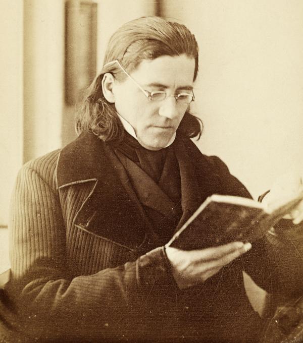 Sepia image of a man with shoulder length hair, wearing spectacles, and holding a book.