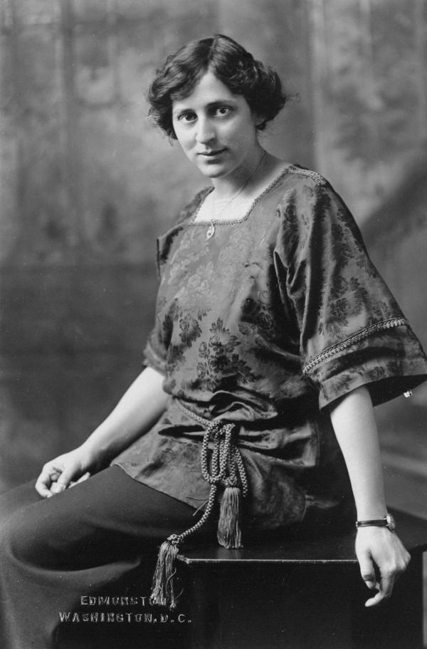 Formal portrait, three-quarter-length, seated, Crystal Eastman Benedict, facing left with upper body and head turned toward camera, wearing square-necked blouse cinched at waist with tasseled-rope belt, watch on left wrist, and necklace.
