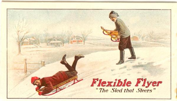 One young boy sleds down a snow covered hill, as another stands at the top awaiting his turn.
