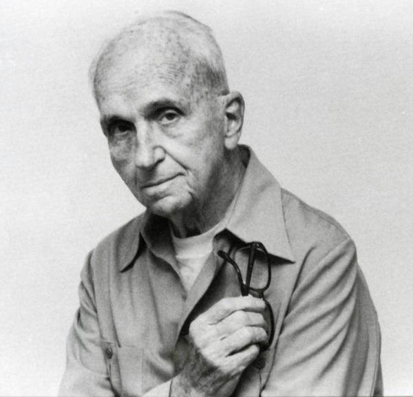 Michener holds a folded pair of glasses against his chest as he poses for an informal, black and white, head and shoulders picture.