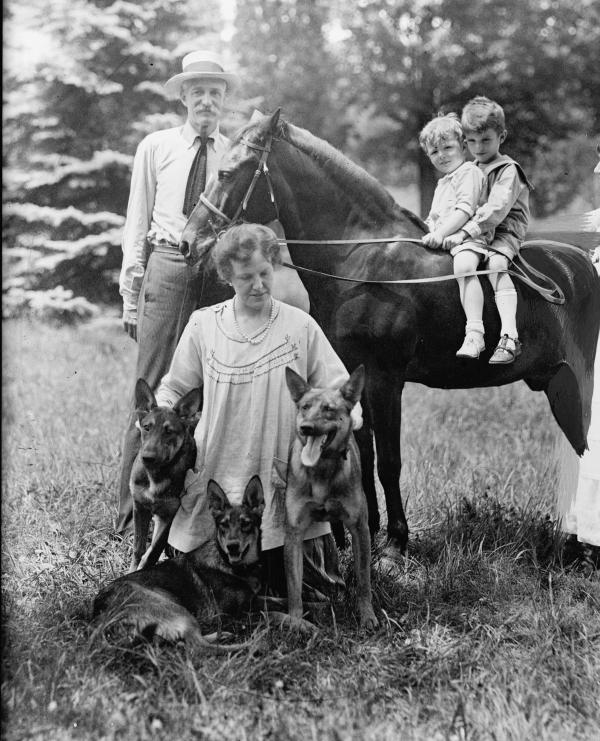Family photo. Two small children sit in a saddle atop a horse. Mrs. Pinchot kneels and pets three german shephard dogs. Pinchot stands at the horse's head.