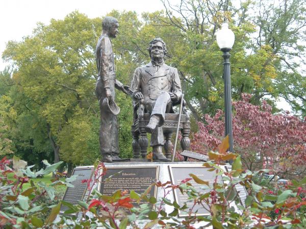 A bronze statue of a man seated and a young boy standing next to him, with one hand outstretched and the other holding a cap. The statue is on a platform and is surrounded by landscaping.