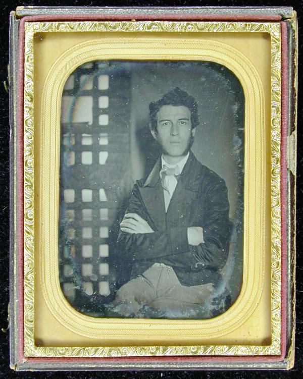 Daguerreotype of a man with mutton chop whiskers, bow tie. Jail door in background.
