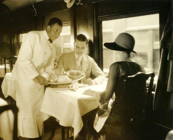 Black waiter dressed in a crisp, white uniform serves two well dressed, white passengers. 