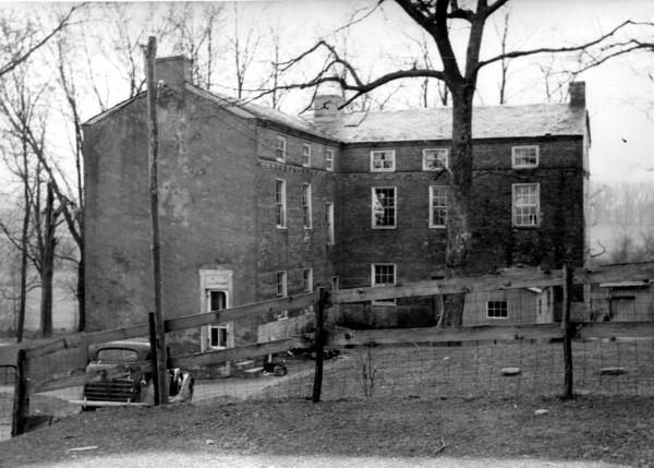 Winter scene of the rear view of Hopewell Academy, A "V" shaped large brick building with a car in the yard.  There is a picket fence in foreground. 