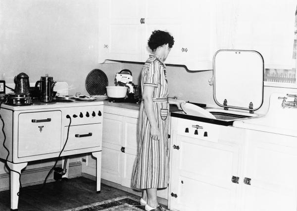 Black and white image of a woman in a striped dress standing at a modernized kitchen sink. A stove is directly behind her and small appliances sit on the counter.