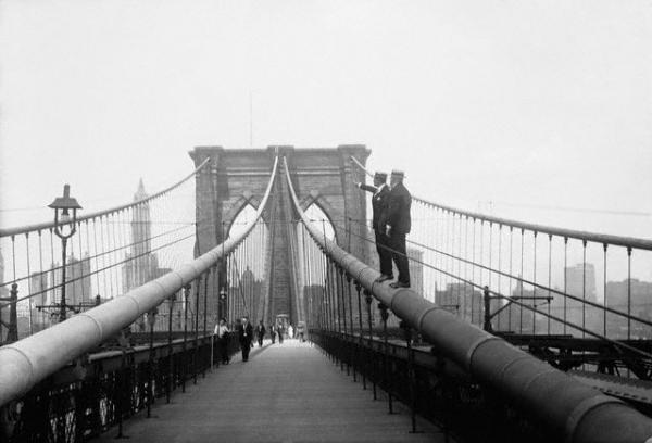 Two men–inspectors–stand upon one of the four cables of the Brooklyn Bridge. One is pointing to a cable which has slipped. New York skyline in background.