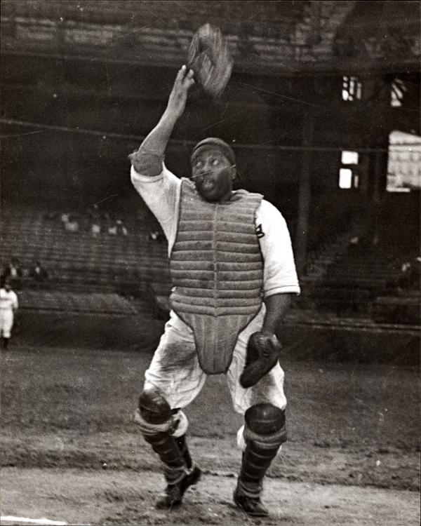 Here Josh Gibson, the 'Black Babe Ruth', famous Negro League baseball player, wearing a white home uniform covered with dirt, and catchers' gear, throws his mask up into the air. 