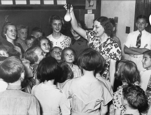Black and white image of a  teacher holding a light bulb in her hand, while surrounded by her amazed students 