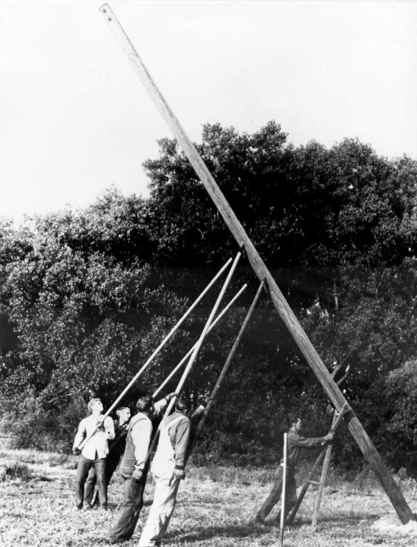 Black and white image of workers installing poles for electrical lines. Pole is slanted in mid installation.