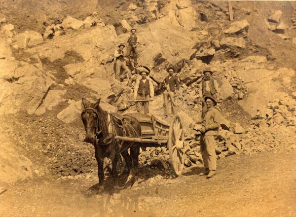One worker stands beside a mule and an Ore Wagon in the foreground of this sepia photograph, as five other male workers stand at different levels on the hillside, holding their tools. 