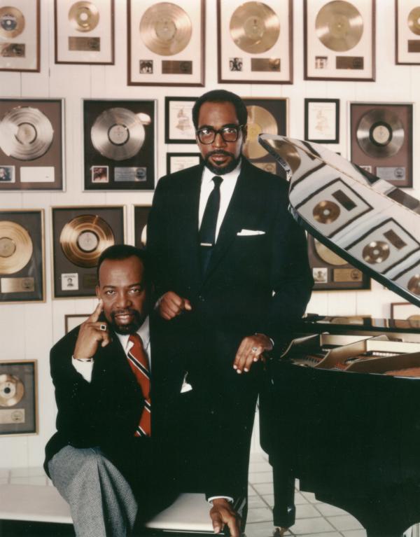 Color photograph of Kenny Gamble and Leon Huff at the piano, with the wall of gold  albums in the background. Reflections of Gold records can be seen in the top of the piano.
