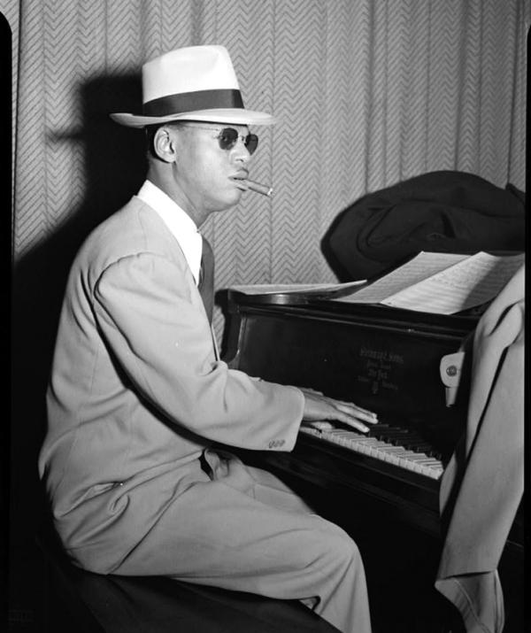 Image of Earl Hines playing the piano. He is wearing a suit, hat, and dark sunglasses.   