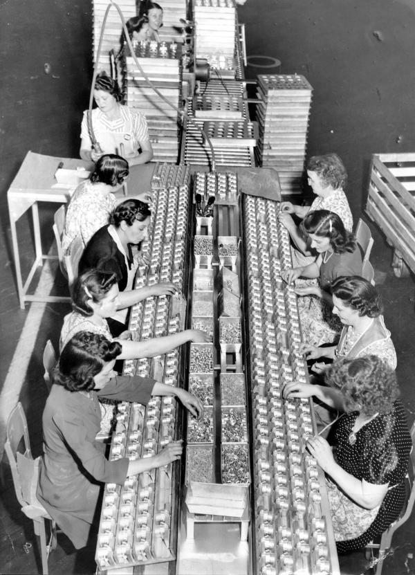 Two rows of women, sit on opposite sides of a huge table as they work on an assembly line.