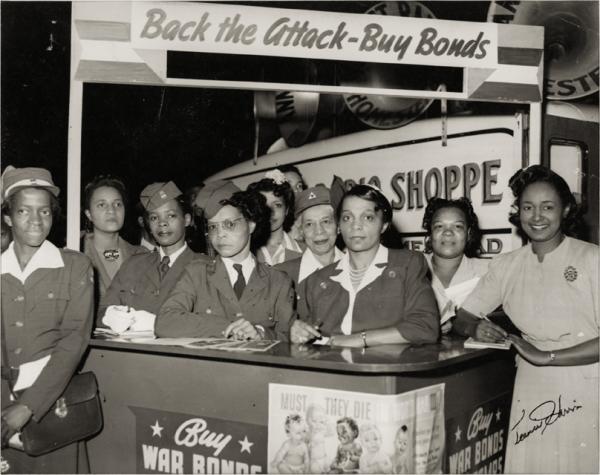  African American women dressed in military clothing stand behind a booth selling war bonds. On the far right is Ruth Gwynnon, who organized this campaign.