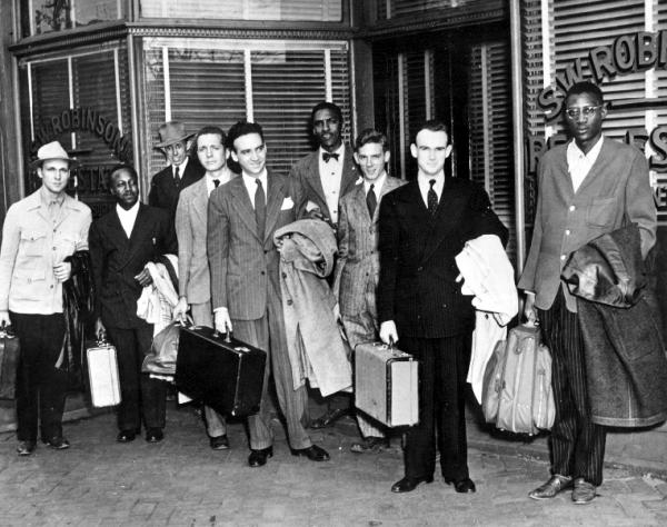 Bayard Rustin (fourth from right) stands with a group of people, some holding baggage, posing for a departure photo.