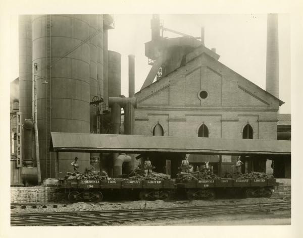 Image of Iron works cast house in the background, two men in business suits appear to be directing the loading of materials by African American workers into Robesonia Iron Company Limited railcars. 