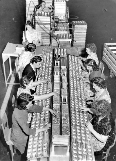 Two rows of women, sit on opposite sides of a huge table as they work on an assembly line.