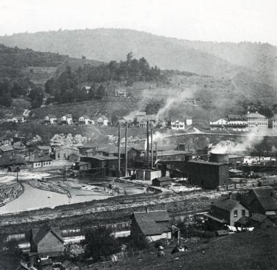 Distant view of the lumbering buidlings and logs in the water of the town. In the center of the photograph one can see the factory with billowing smoke rising in to the air and along the outskirts sit the houses.  
