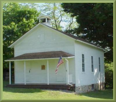 Exterior view of white one-room school house complete with flag and a bell.