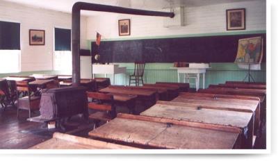 Interior, room with desks, a blackboard stretches across the back wall, and  a large black stove with a pipe extending to annd across the ceiling.  