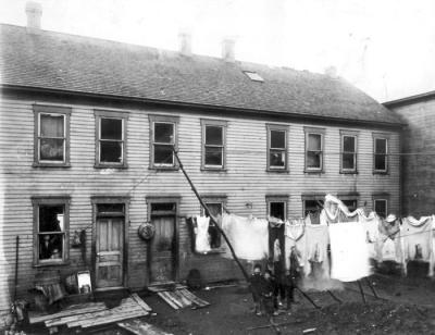 In this backyard there are several lines of laundry hanging from lines that are attached to the house and extend to poles. Several small children pose underneath one of the lines for this photograph. The house is a long, two- story, wooden, structure with eight windows across the top floor. On the bottom floor there is a window to the left and two small children peer out from the inside. To the right of this window are two doors, with a wash tub hanging between the two. To the right of the doors, five windows fill the rest of the bottom floor walls. The yard is filled with debrie.