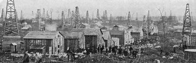 A group of hastily constructed buildings stand in a line along a primitive road in front of a field full of oil derricks.