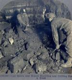 Black and white photograph showing two miners shoveling and drilling for a blast.
