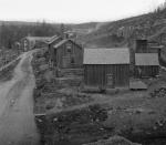 Black and white photograph of a dirt road lined with small, stark houses. In the background sits a large building.
