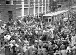 A large crowd forms in a public street surrounding cable cars and stopping  traffic.