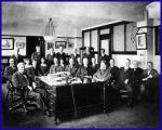 A large group of men sitting at and standing around a desk, posing for a group photograph.