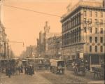 The intersection; horse-drawn carts and wagons, streetcars and tracks, and people in the street. Stores line the street, decorated with advertising, including: <i>Tower Hall</i>; <i>Garitee and Son</i>; <i>American Clothing Co.</i>; <i>Williamson and Cassedy</i>; <i>Oakhall</i>; <i>Wanamakerand Brown.</i>  